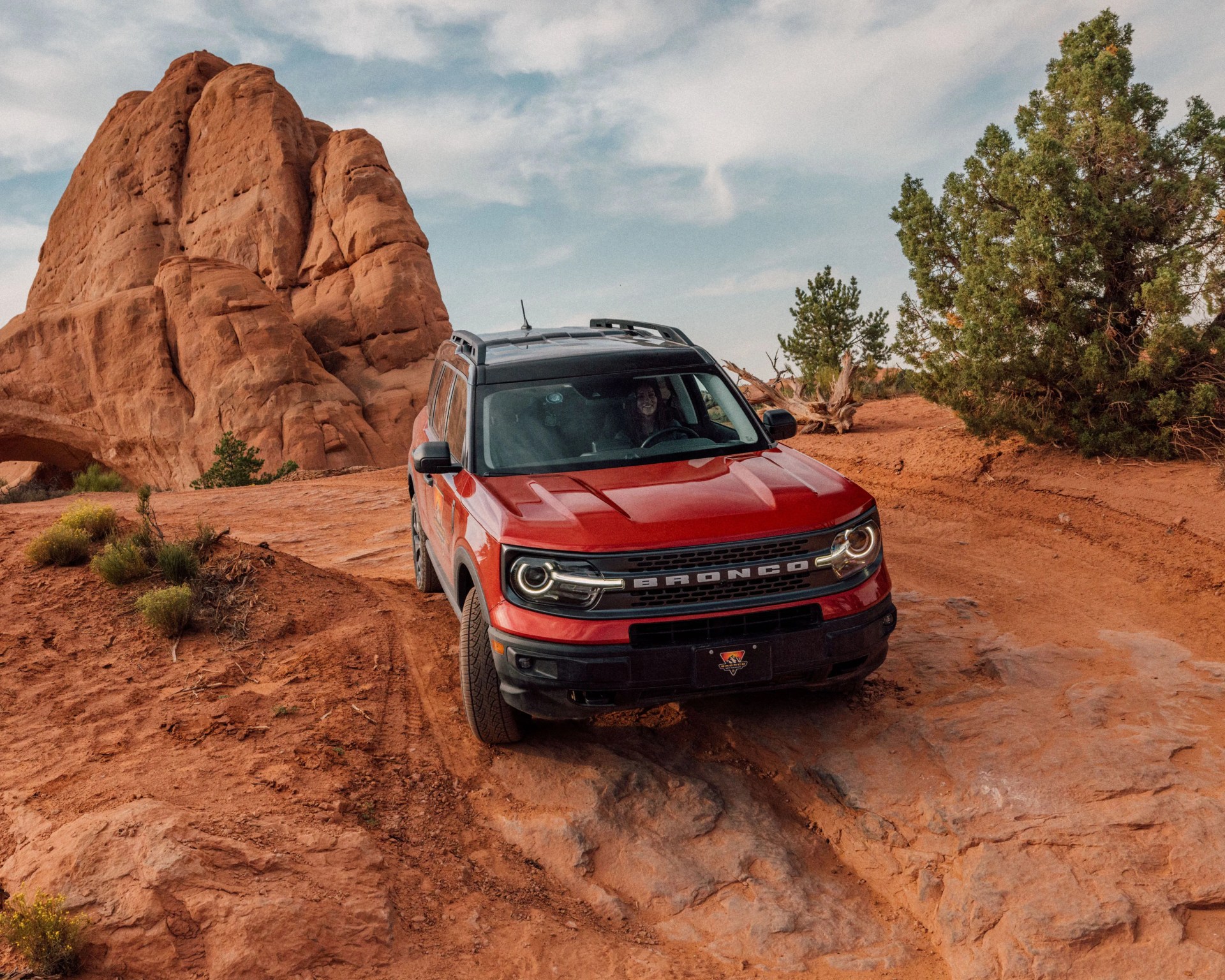 Red Ford Bronco SUV on rocky desert terrain near large sandstone formation and green bushes.