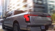 Silver Ford F-150 Lightning pickup truck driving on a city street with illuminated rear lights.