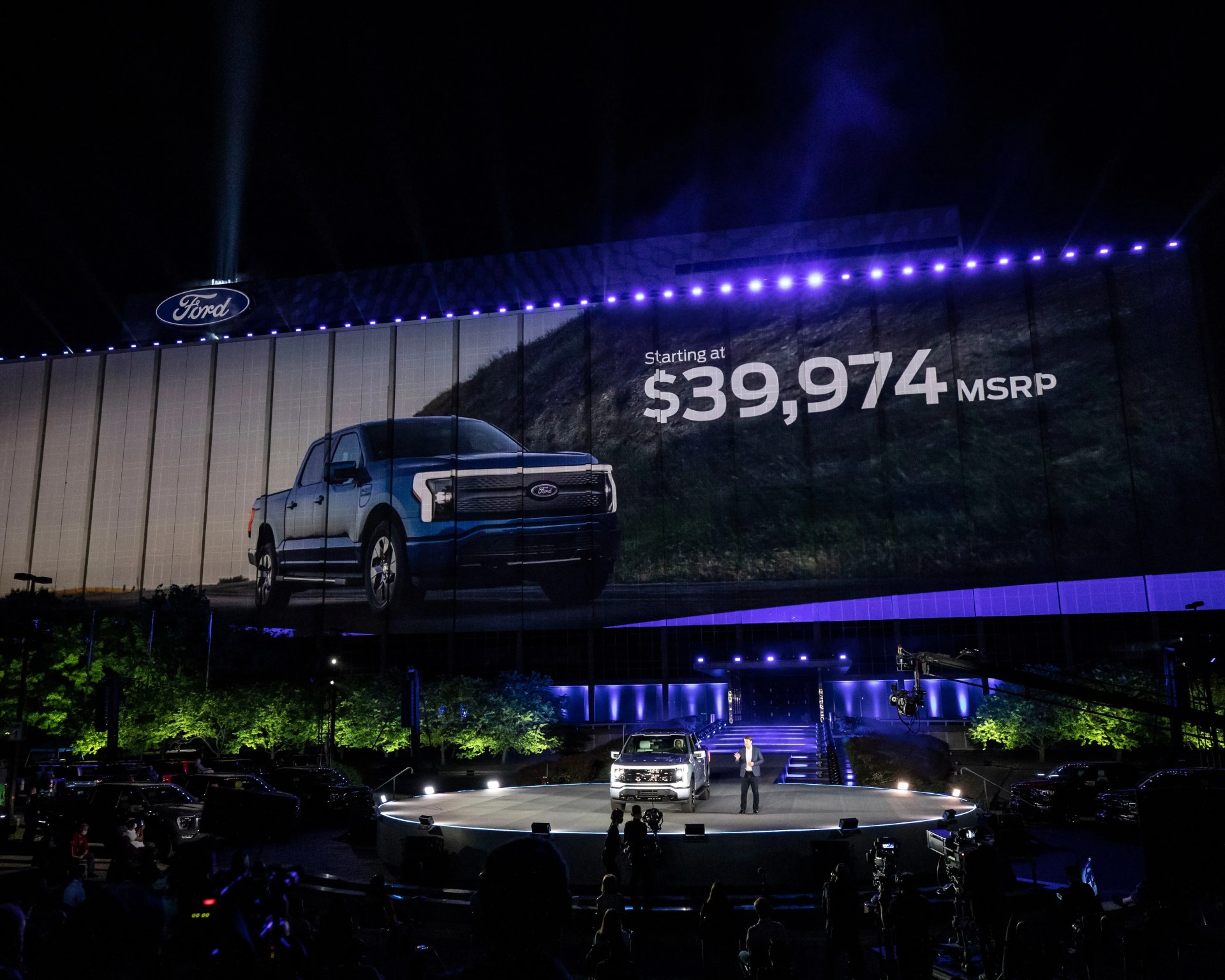 Large outdoor Ford advertisement showing a blue pickup truck and price starting at $39,974 MSRP above a stage with a person and a white truck.