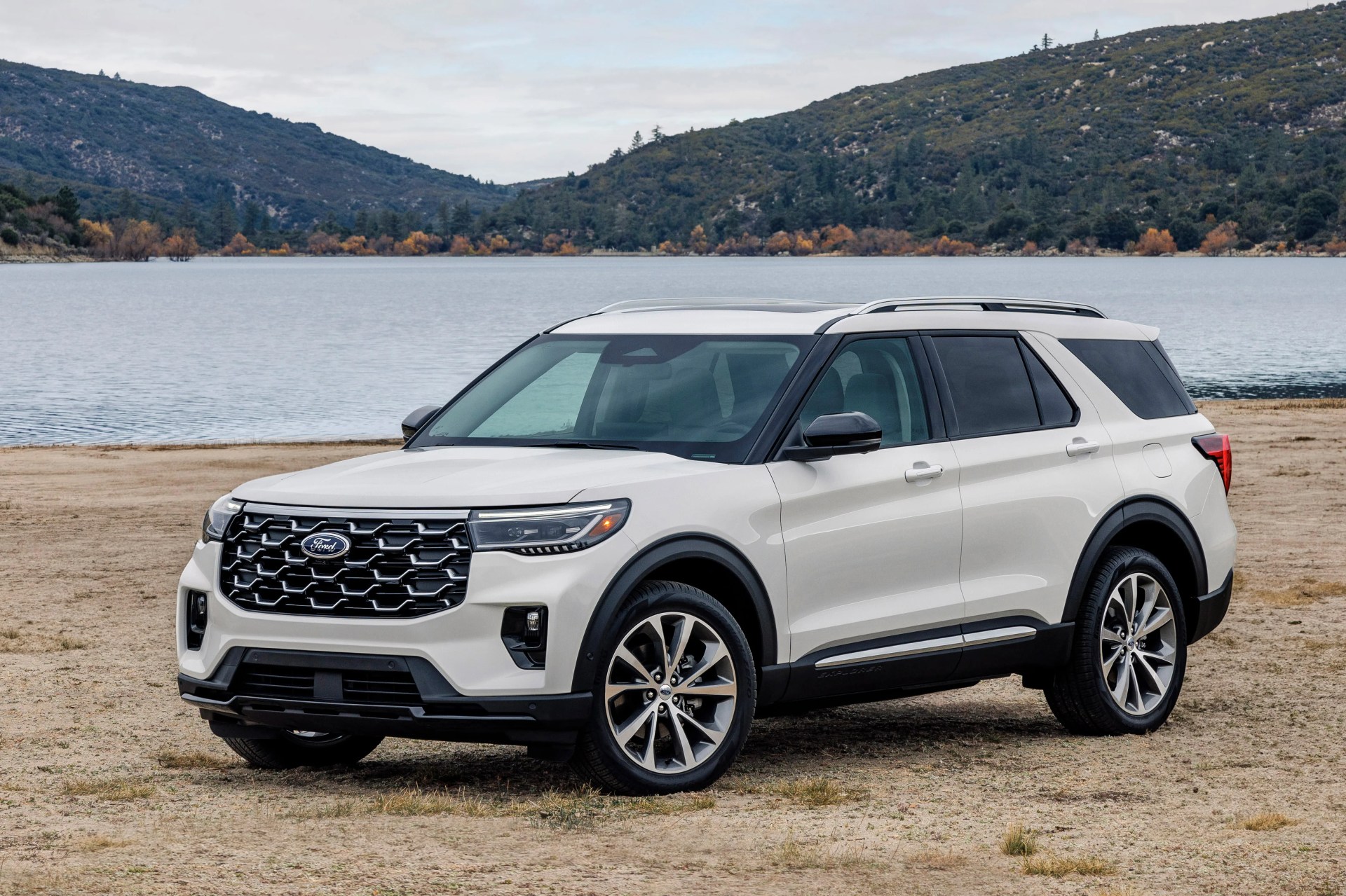White Ford Explorer SUV parked on dry ground near a lake with forested hills in the background.
