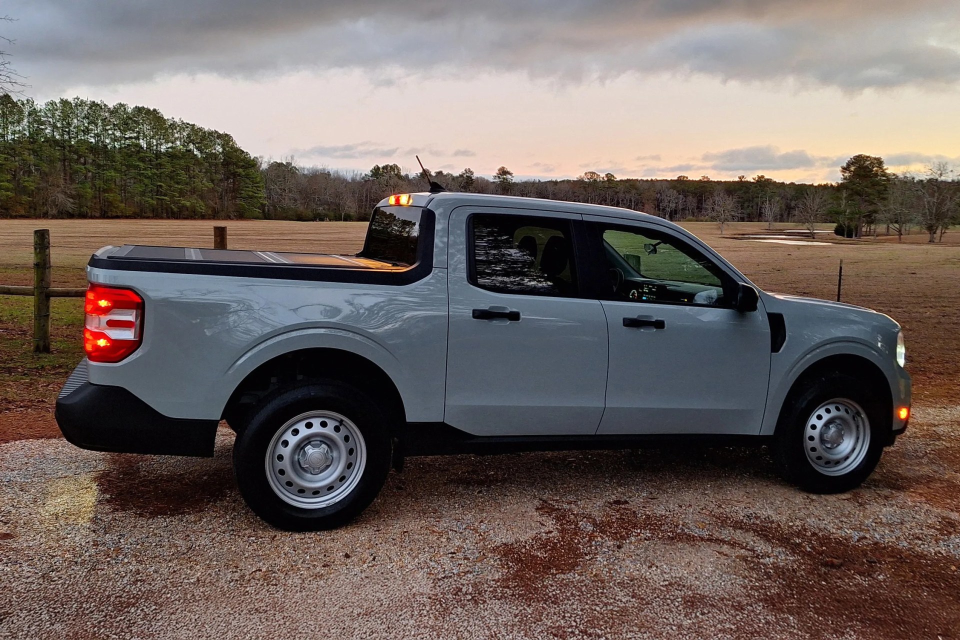 Light gray pickup truck with black trim and steel wheels parked on gravel near a field at dusk.