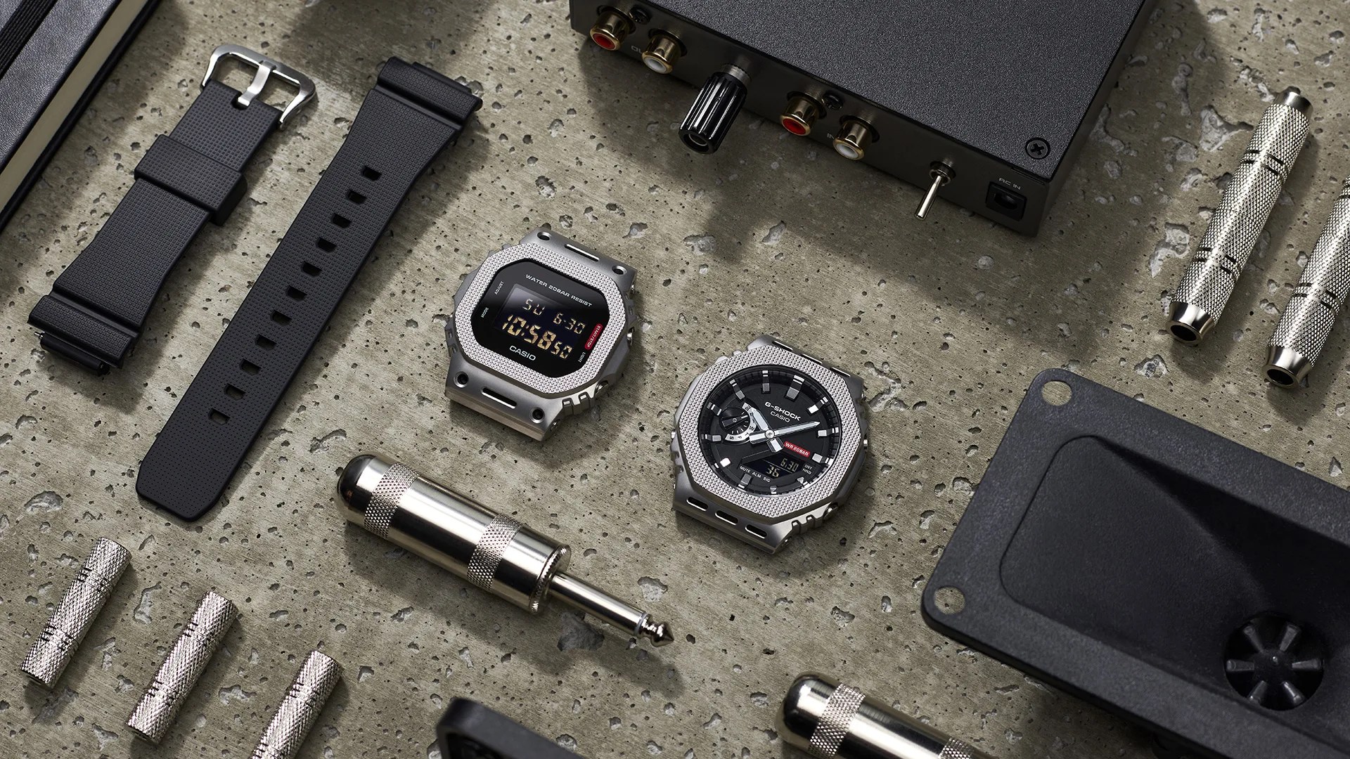 Two Casio G-Shock watches with silver octagonal bezels and black faces, one digital and one analog-digital, on a textured gray surface with black watch straps and audio connectors.