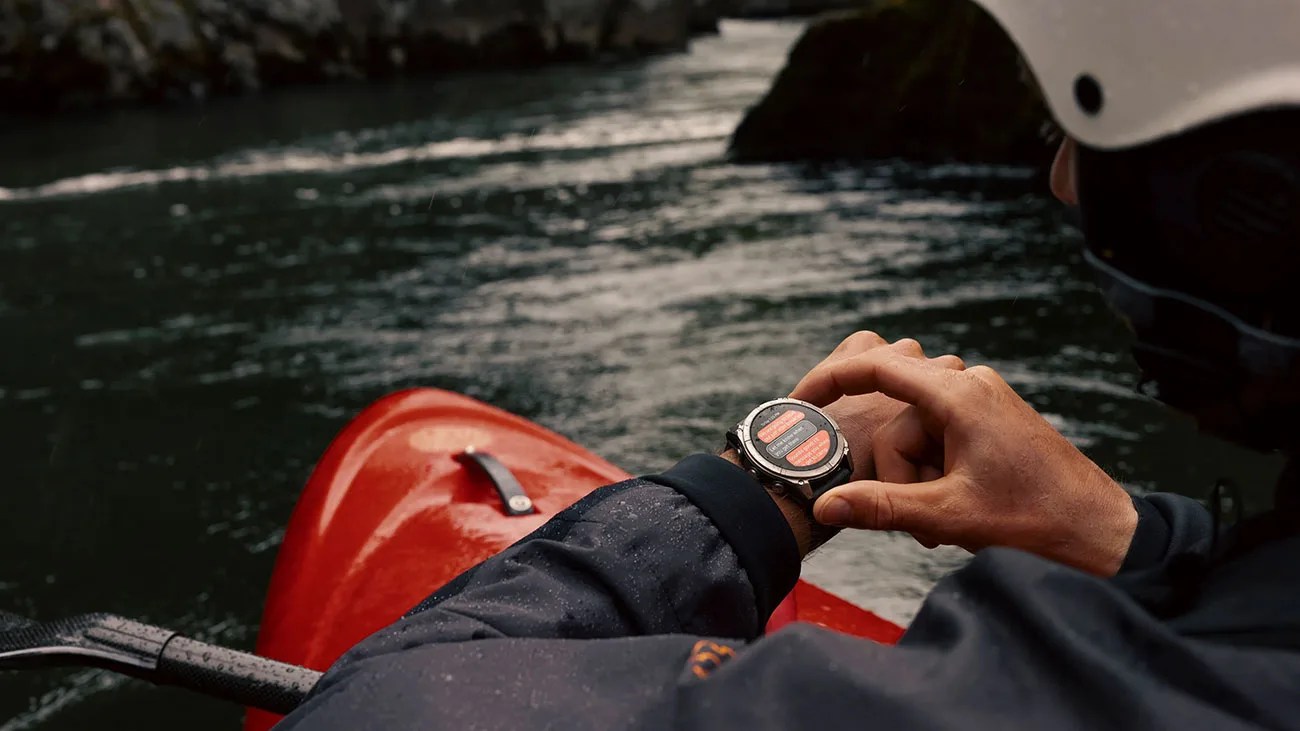 Person wearing a helmet and dark jacket checking a smartwatch while kayaking on a river.