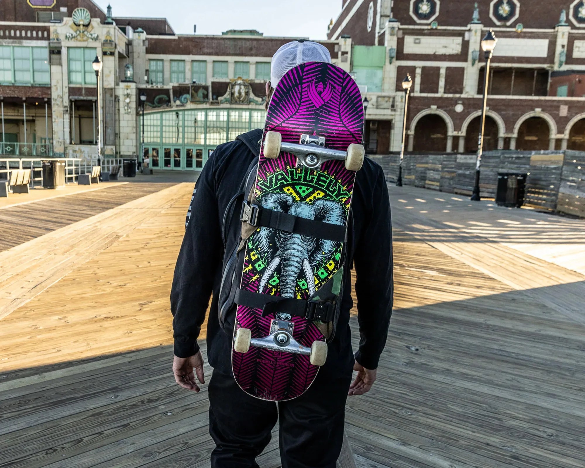 Person wearing a black hoodie and white cap carrying a skateboard with an elephant graphic on a wooden boardwalk.