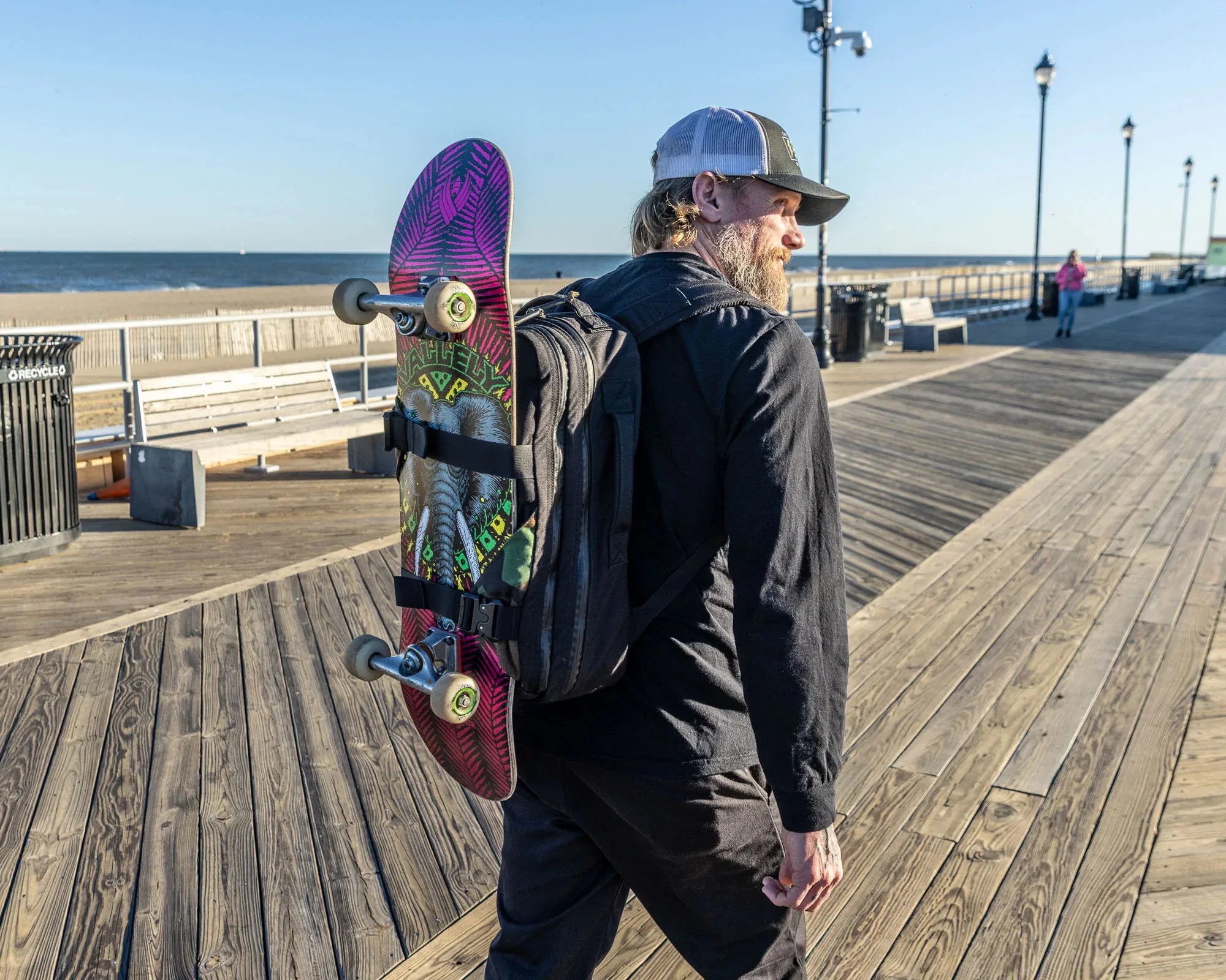 Man wearing a black backpack with a skateboard featuring an elephant graphic strapped to it on a wooden boardwalk by the beach.