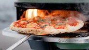 Wood-fired pizza with basil and pepperoni being removed from a Gozney oven with a metal peel.