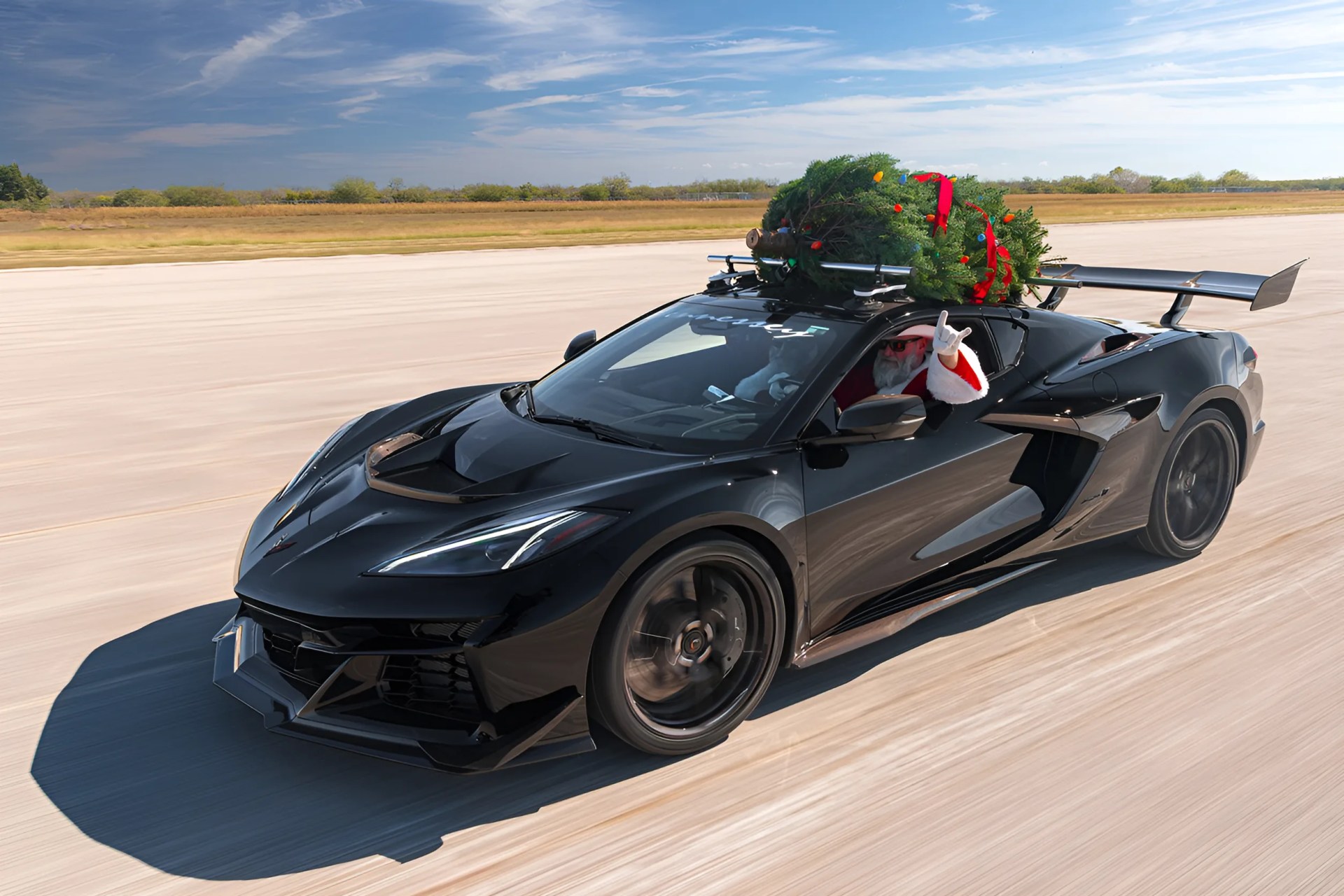 Black sports car with a large rear spoiler carrying a decorated Christmas tree on the roof, driven by a person in a Santa suit.