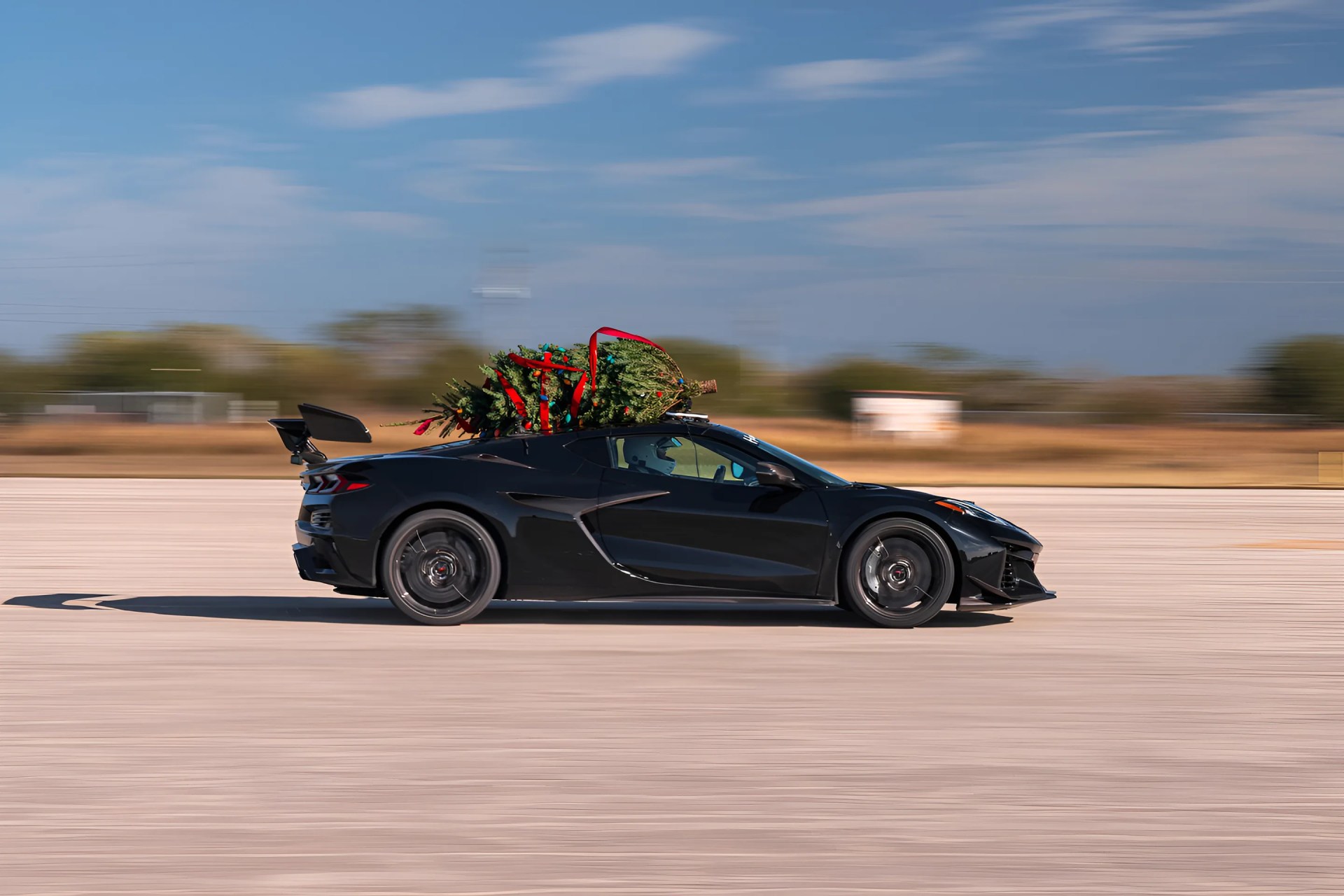 Black sports car driving on a road with a decorated Christmas tree tied to its roof.