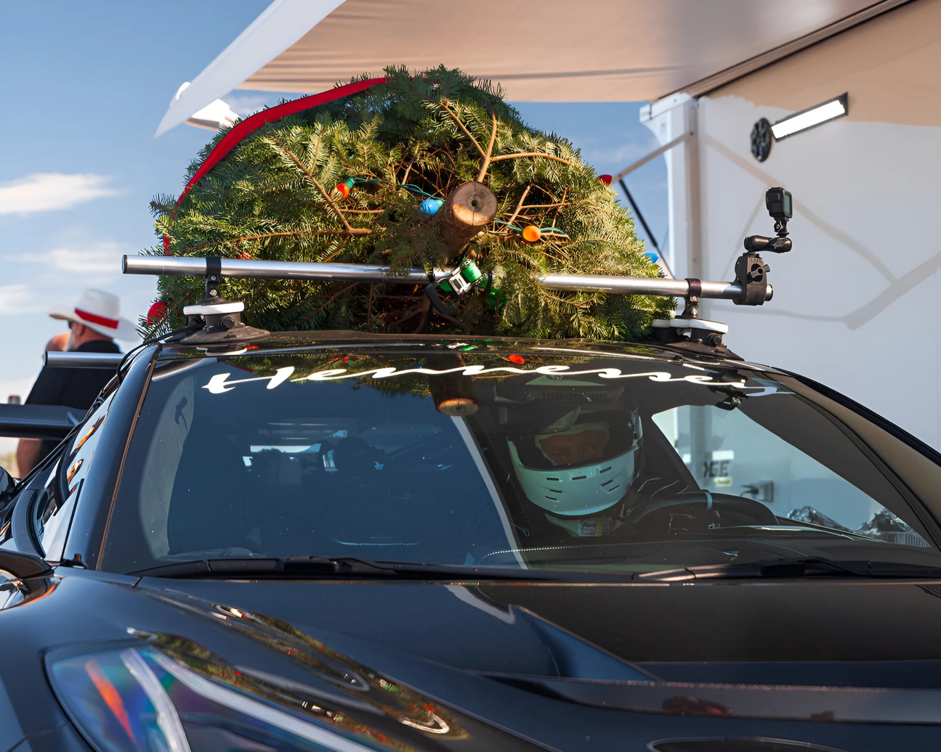 Black sports car with a Christmas tree secured on the roof rack and a driver wearing a white helmet inside.