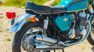 Close-up of the rear side of a vintage blue Honda motorcycle showing the exhaust pipes, rear tire, and seat.