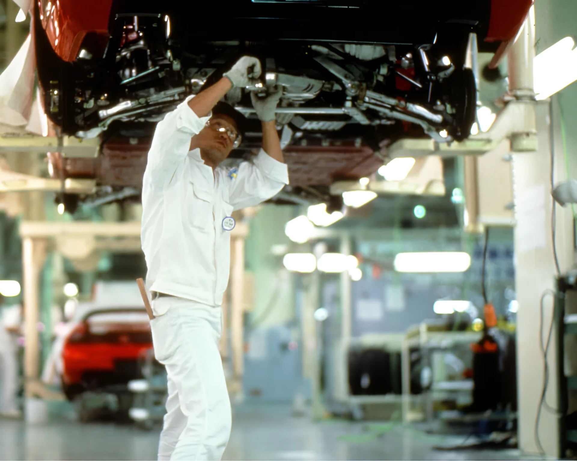 Mechanic in white uniform working under a raised red car in a brightly lit auto repair shop.