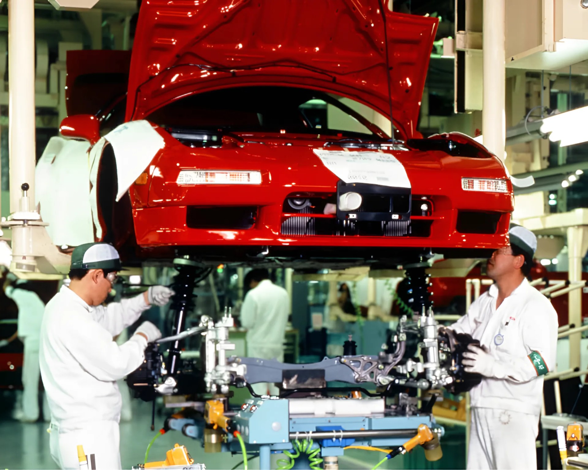 Red sports car with hood open suspended above two workers assembling parts in a factory setting.