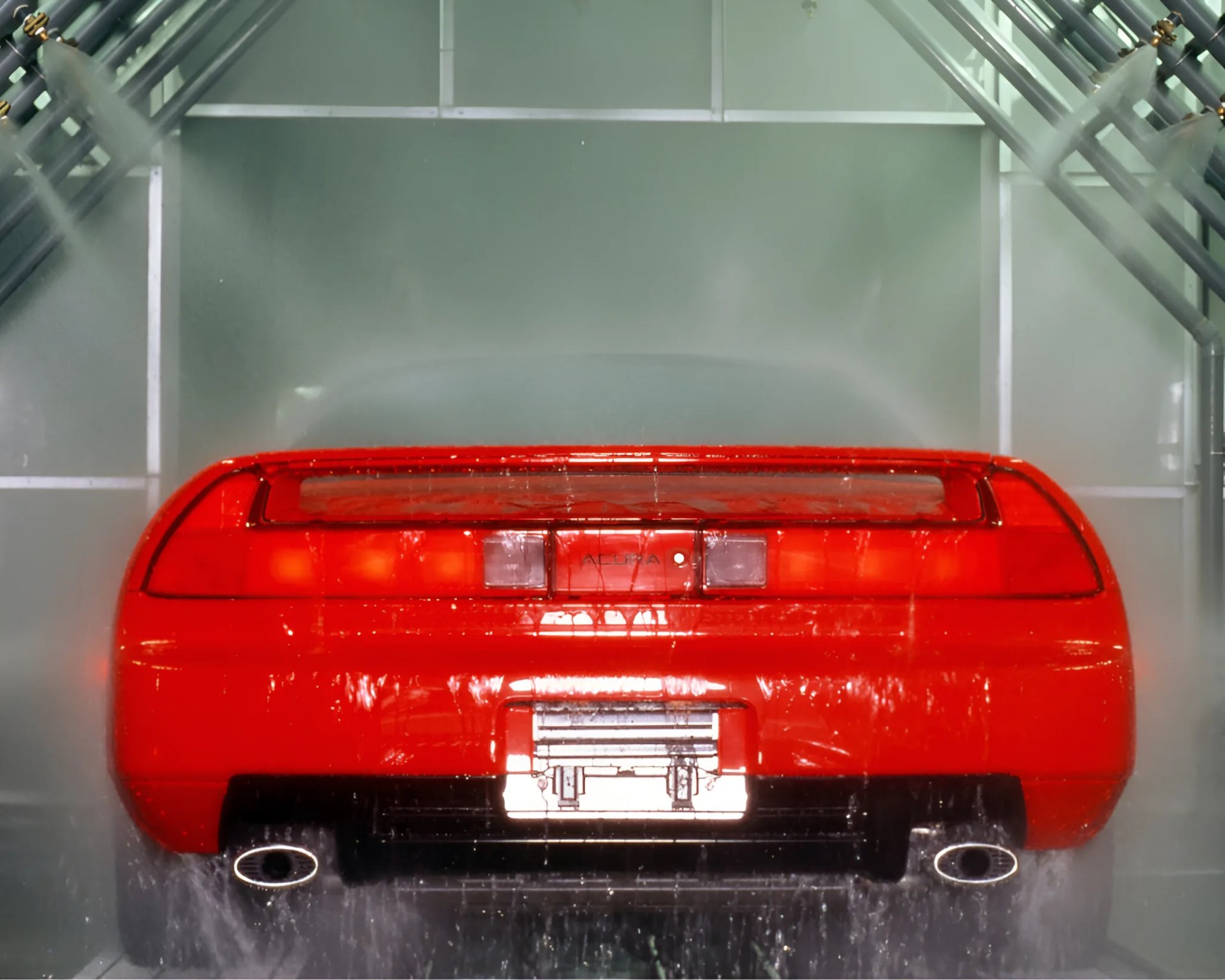 Rear view of a red sports car with dual exhausts in a water spray testing chamber.