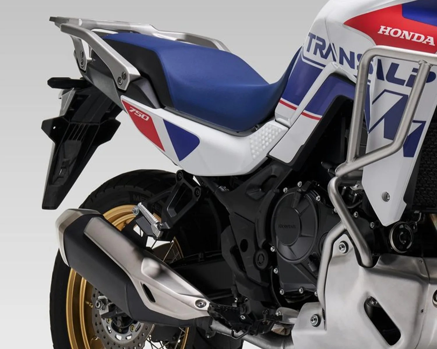 Close-up of the rear section of a white, blue, and red Honda Transalp 750 motorcycle with a blue seat and gold wheel.