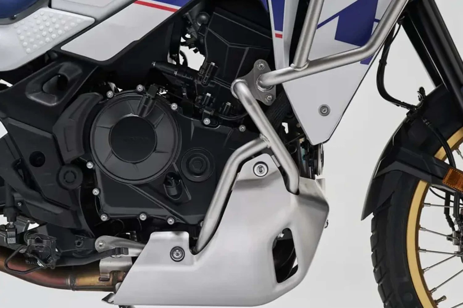 Close-up of a motorcycle engine with a black casing, silver crash bars, and a silver skid plate, showing part of the front wheel with a gold rim.