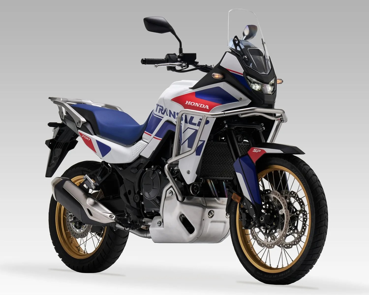 White, blue, and red Honda Transalp 750 adventure motorcycle with gold rims and protective crash bars.