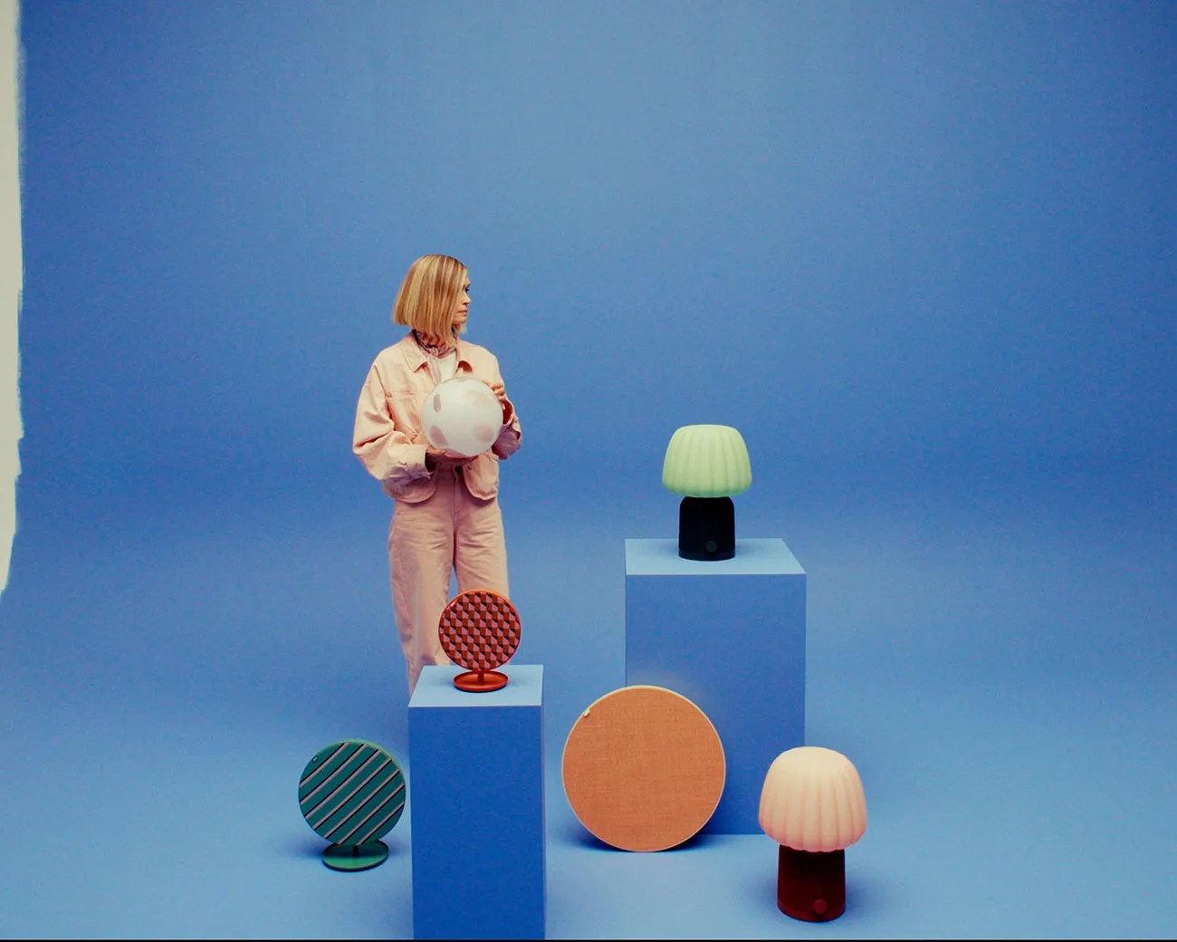 Person in a light pink outfit holding a white ball with pink spots, surrounded by colorful round speakers and lamps on blue pedestals.