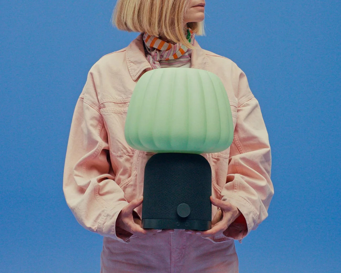 Person holding a black speaker with a large green ribbed lampshade on top against a blue background.