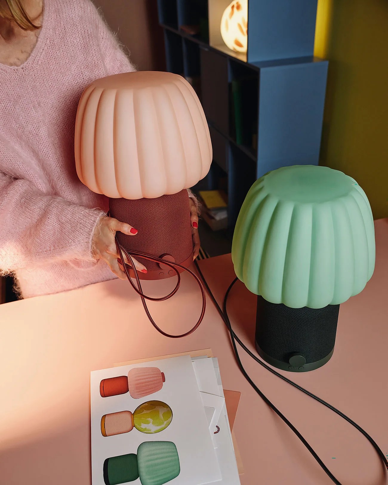 Two mushroom-shaped table lamps with fabric bases, one pink and one green, on a pink surface with a person holding the pink lamp.