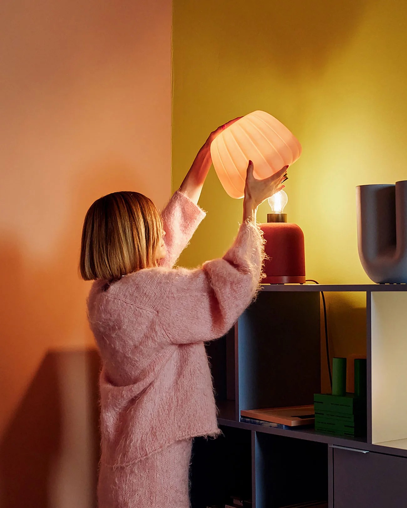 Person in a pink fuzzy sweater placing a ribbed, glowing lamp shade on a red table lamp on a shelf.