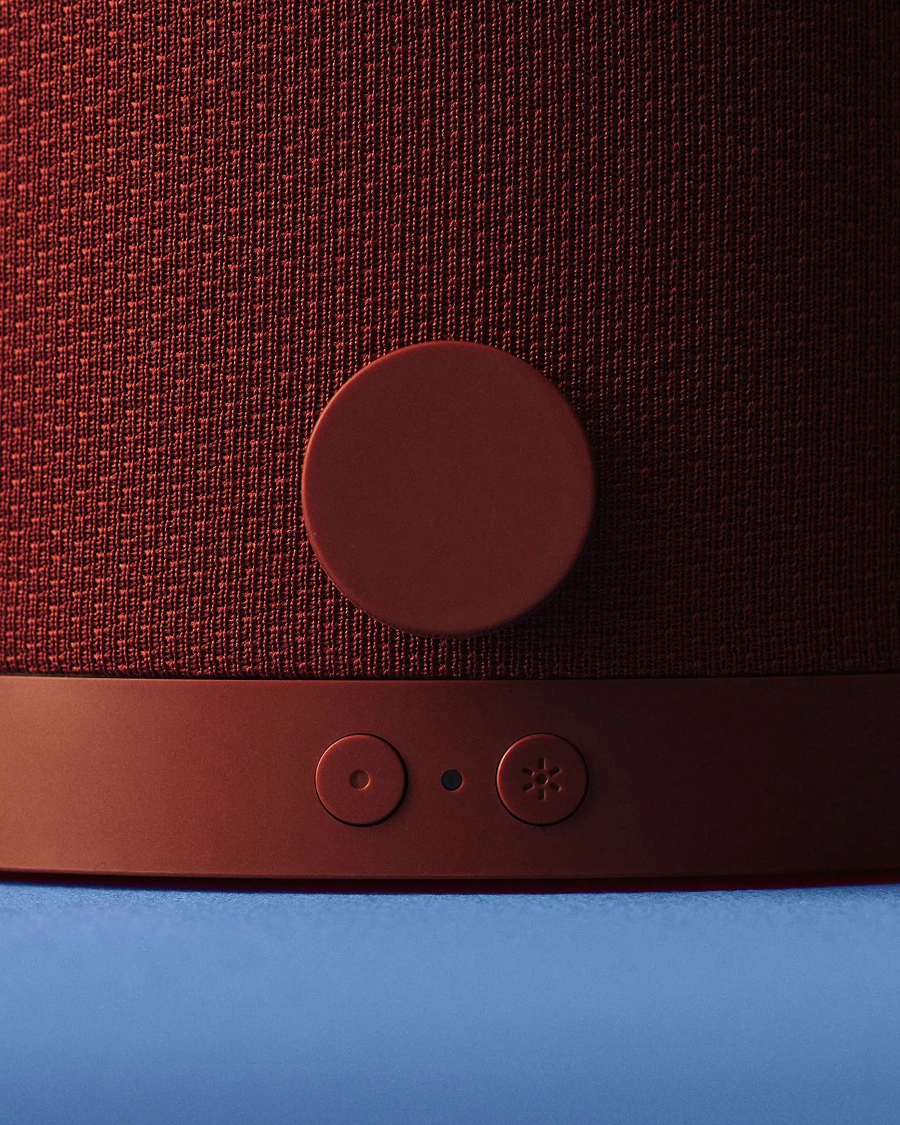 Close-up of a maroon fabric speaker with a large round knob and two smaller buttons on a blue surface.