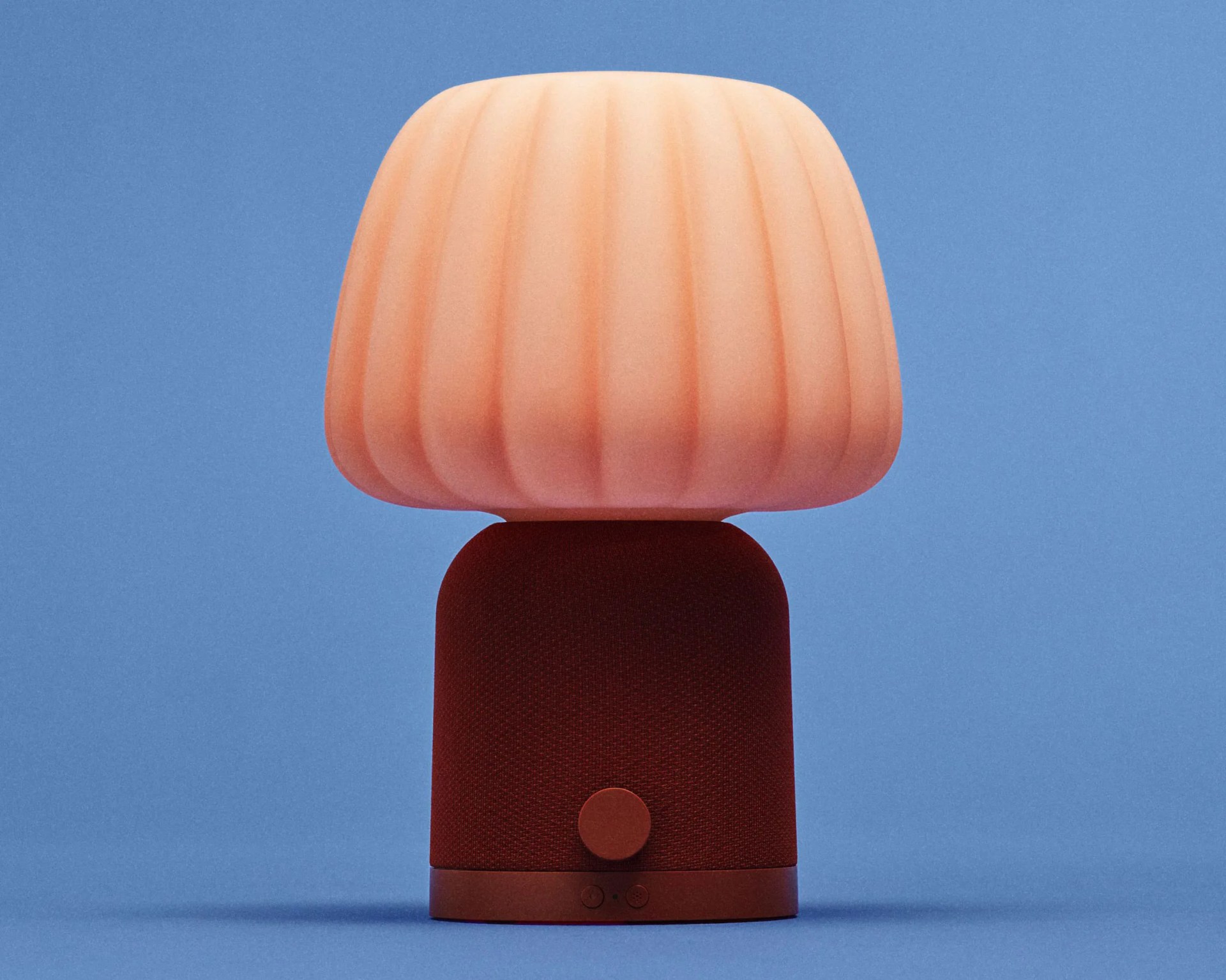 Red fabric-covered smart speaker with a rounded pink ribbed lampshade on top against a blue background.