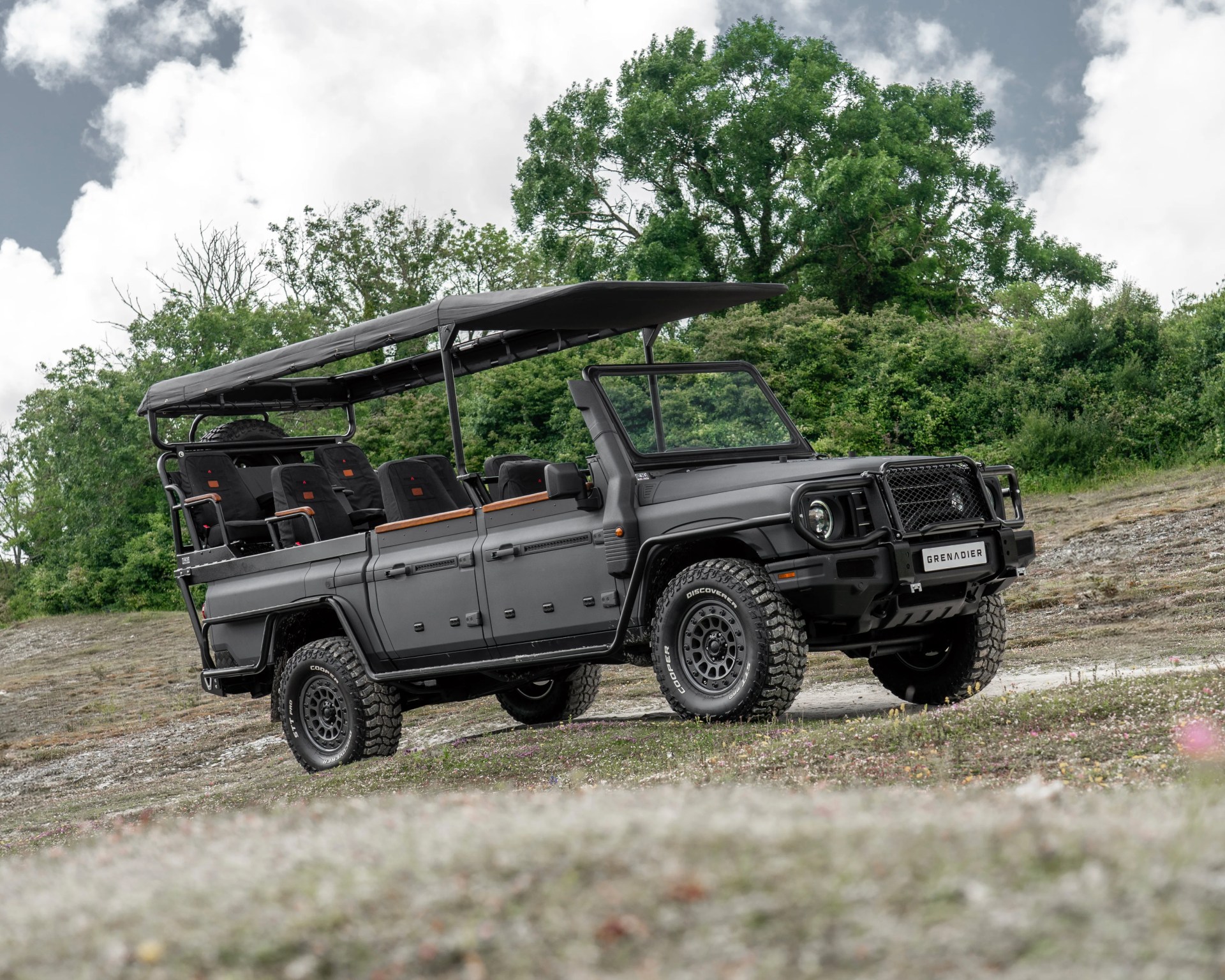 Matte black off-road Grenadier vehicle with open sides, canopy roof, and rugged tires on a grassy slope.