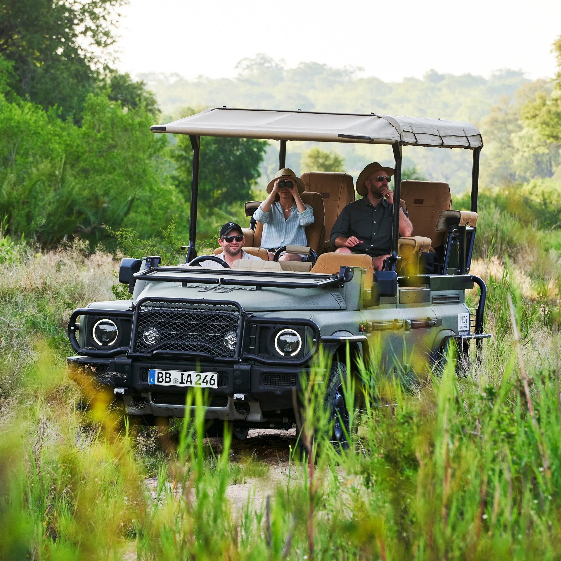 Open-top safari vehicle with tan seats carrying three people, driving through green grass and bushes.