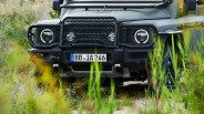 Front view of a matte green off-road vehicle with round LED headlights and a black grille guard, partially obscured by tall grass.