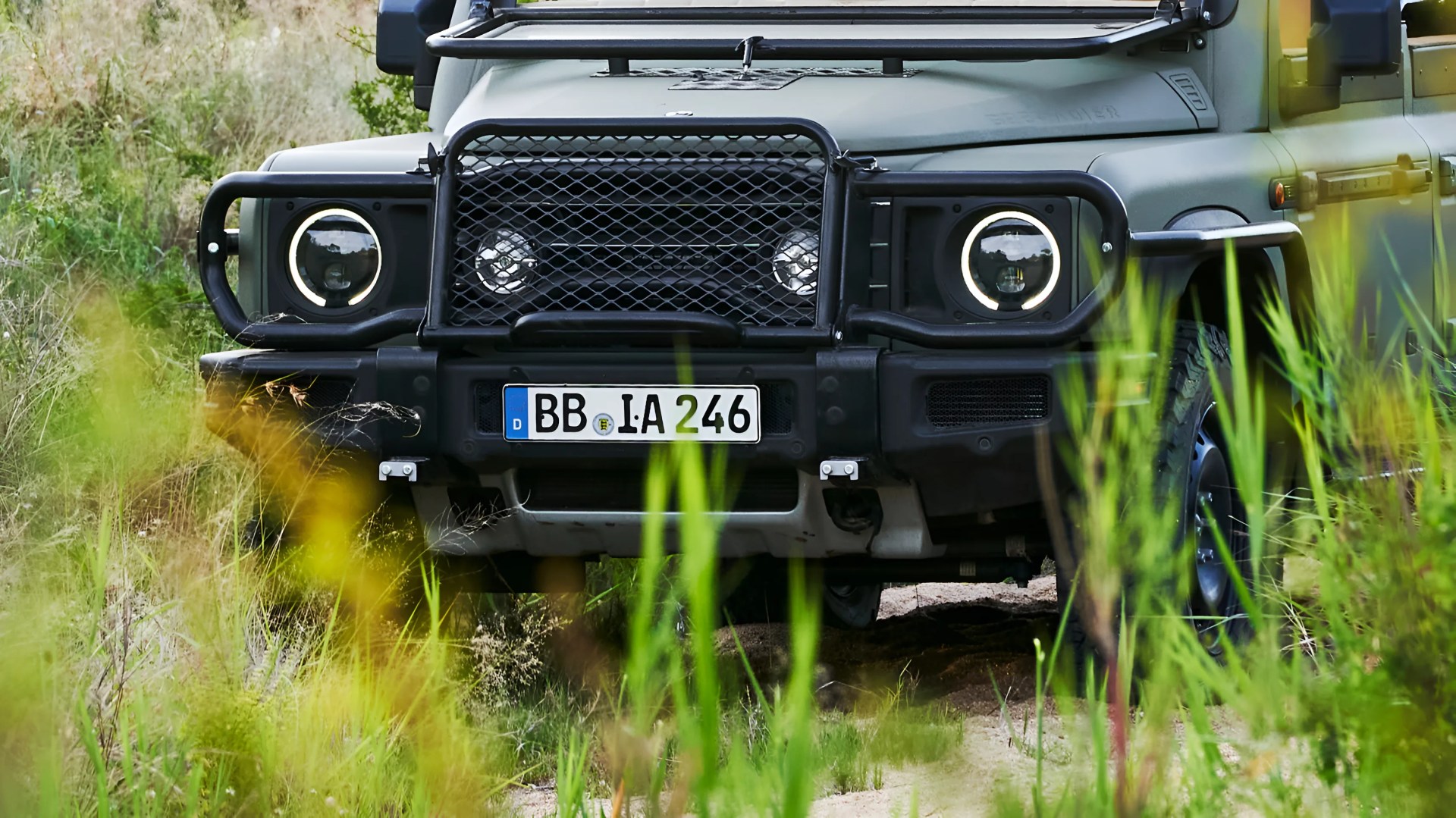 Front view of a matte green off-road vehicle with round LED headlights and a black grille guard, partially obscured by tall grass.