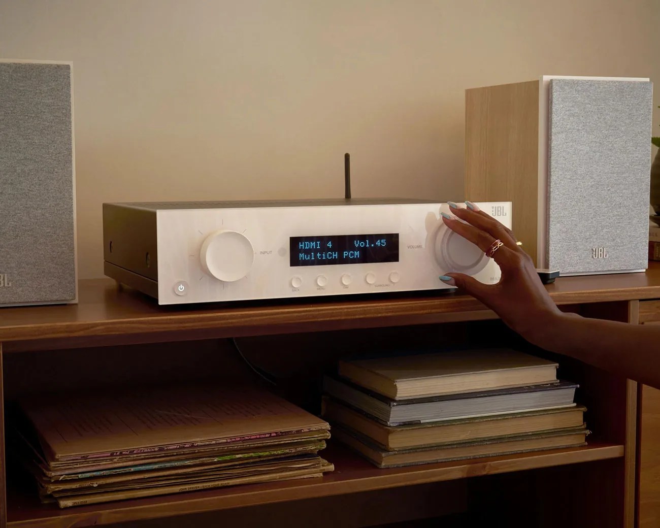 White JBL audio receiver with digital display and two gray fabric-covered speakers on a wooden shelf, hand adjusting volume knob.