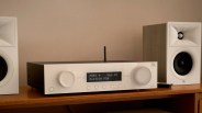 White JBL MA310 audio receiver with black top and digital display showing HDMI 4 and volume 45, flanked by white JBL speakers on a wooden shelf.