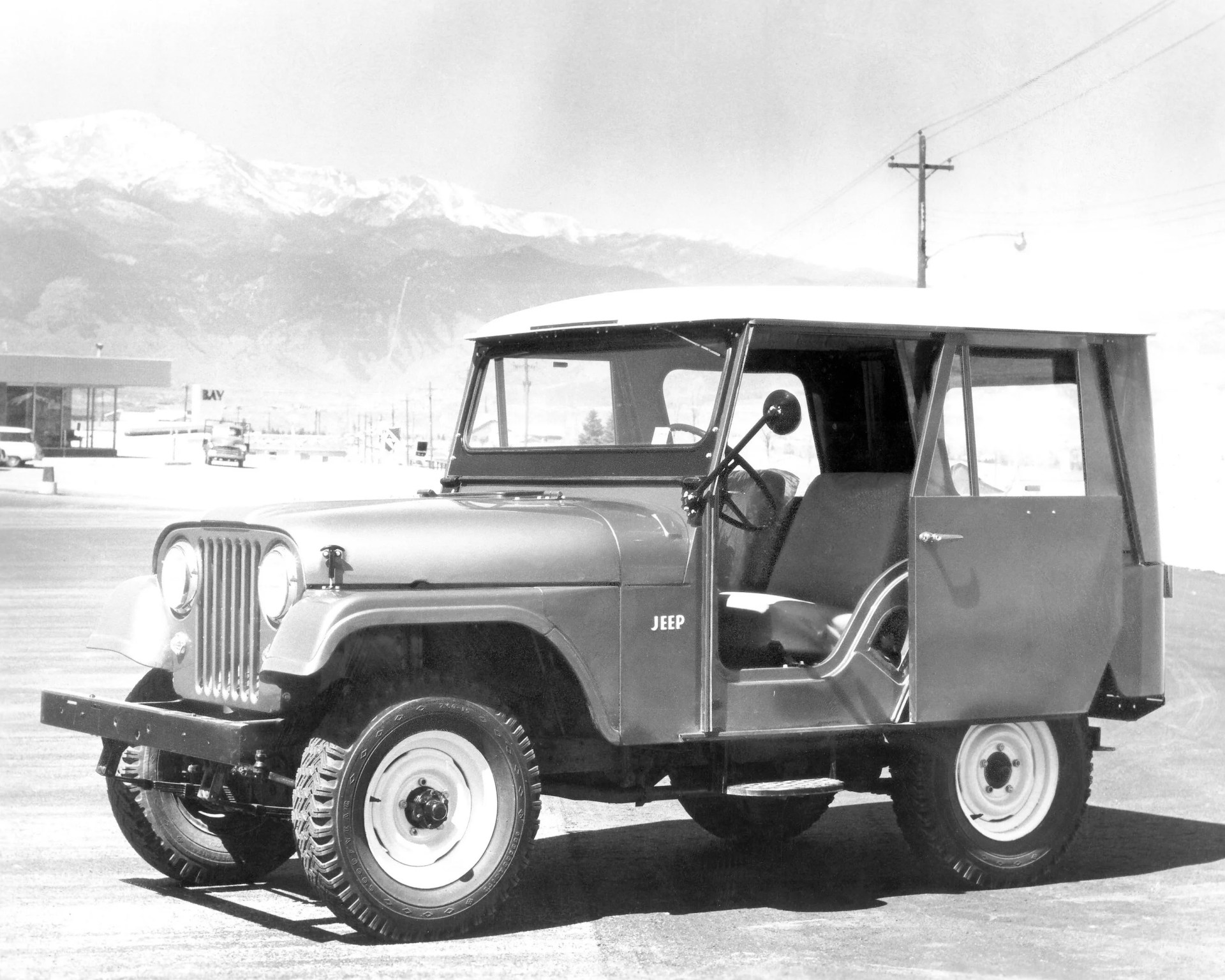 Vintage Jeep with a boxy hardtop, open driver's door, and white steel wheels parked on a paved area with mountains in the background.