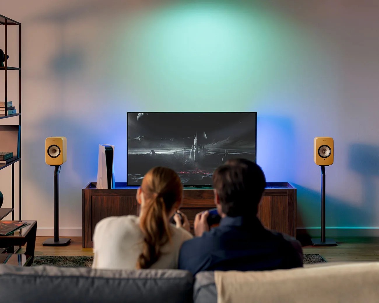 Two people sitting on a couch playing video games on a TV with yellow speakers on stands and a white gaming console on a wooden cabinet.