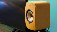 Yellow KEF wireless speaker with a central cone driver and mesh grille on a black stand.