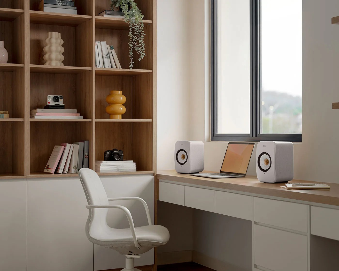 Modern home office with white swivel chair, wooden desk with laptop and two white speakers, and wooden shelving with books and decor.