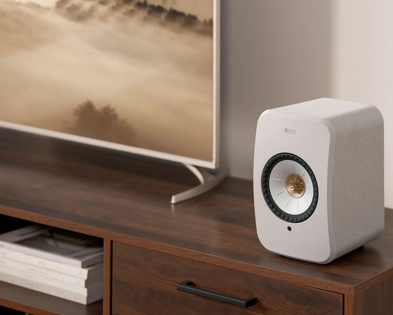 White KEF speaker with gold and silver driver on a wooden media console next to a TV.