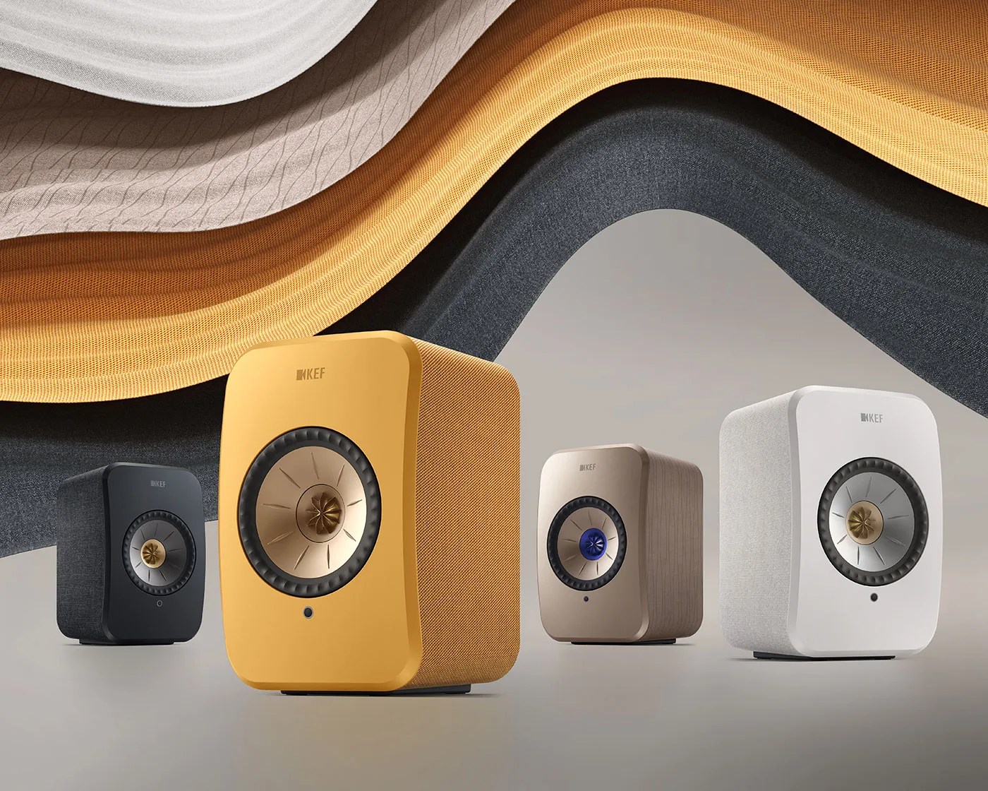 Four KEF speakers in black, yellow, beige, and white with fabric covers and metallic drivers, set against a wavy fabric backdrop.