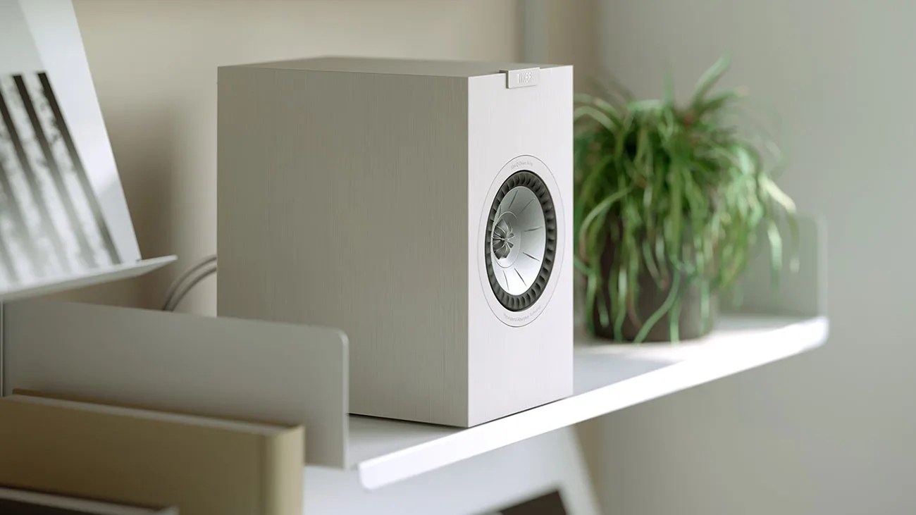 White rectangular KEF speaker with silver driver on a white shelf next to a green potted plant.