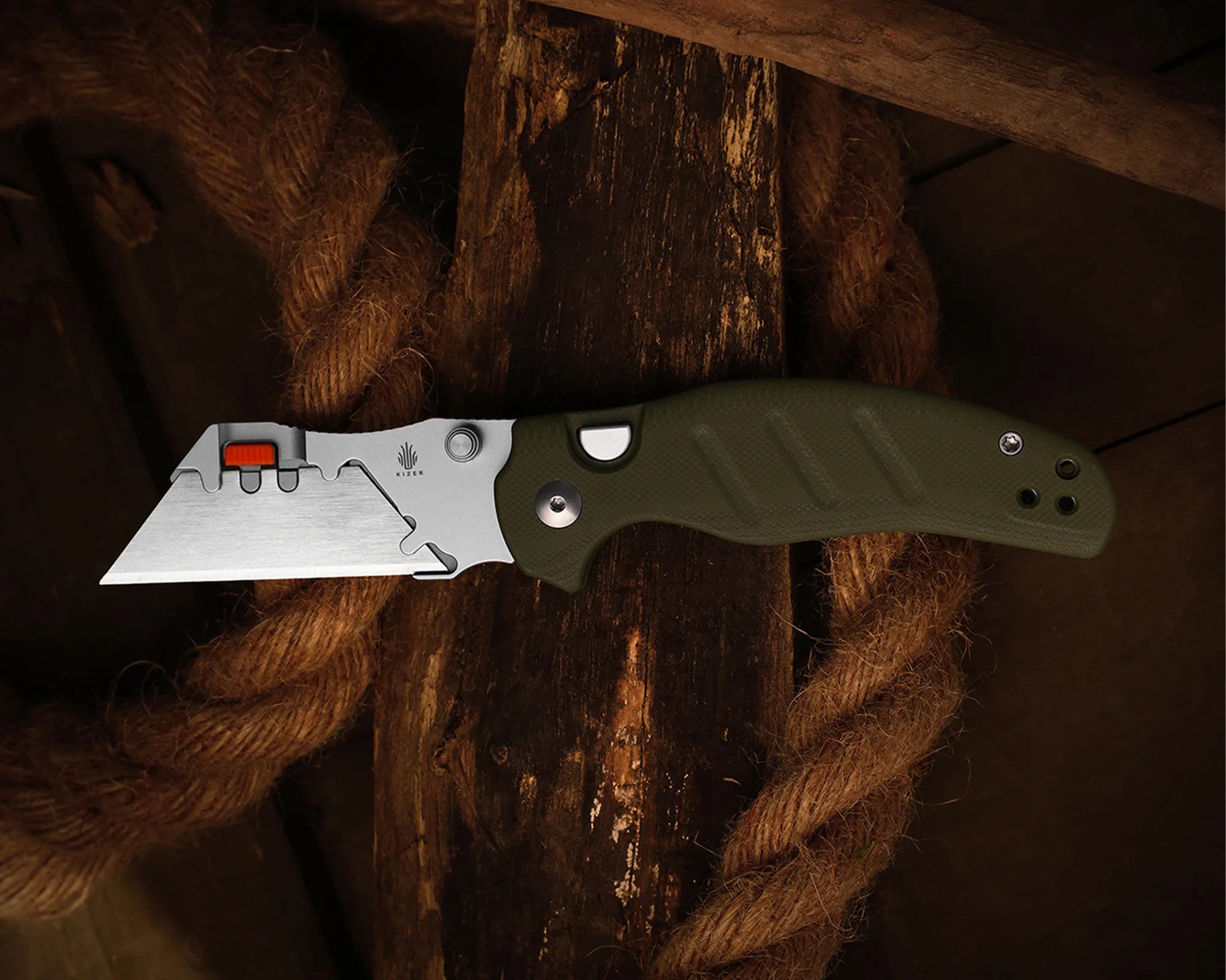 Folding utility knife with a silver blade and olive green textured handle resting on a wooden surface with coiled rope.
