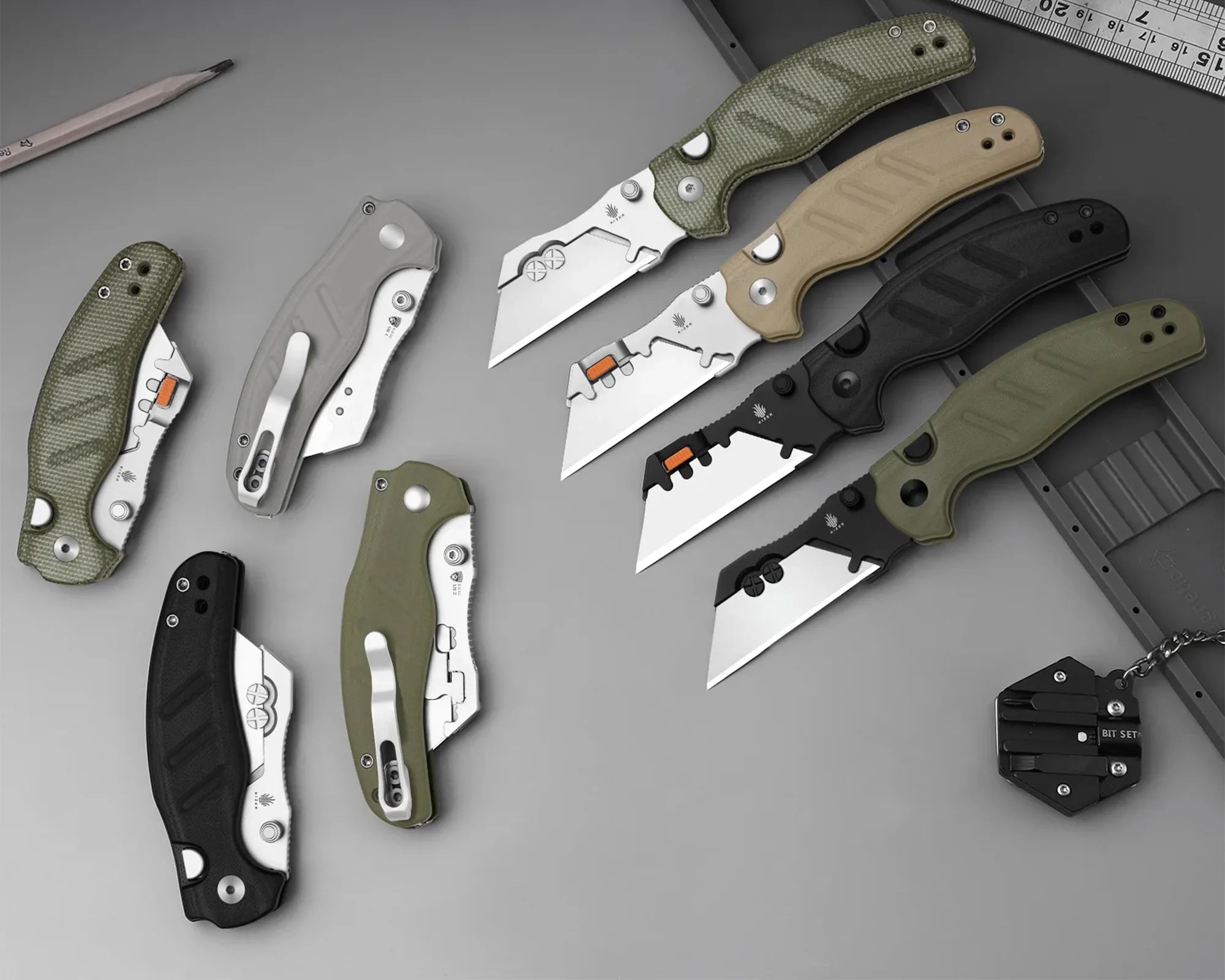 Eight folding utility knives with various handle colors (green, tan, black, gray) and a small black multi-bit tool on a gray surface.