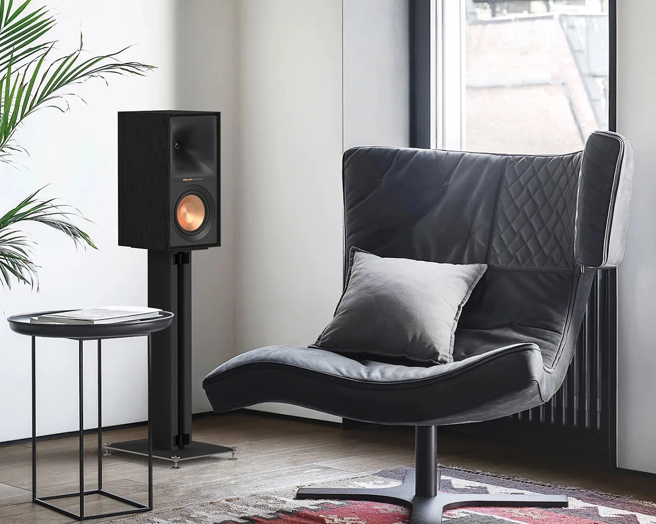 Black leather lounge chair with a gray pillow next to a black speaker on a stand and a small round black side table.