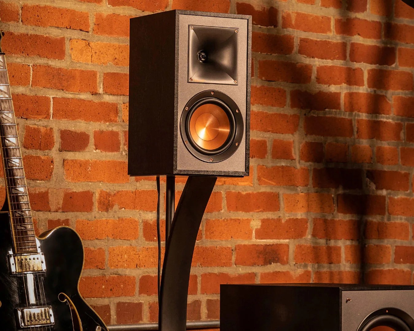 Black bookshelf speaker with copper woofer on a curved black stand against a brick wall.