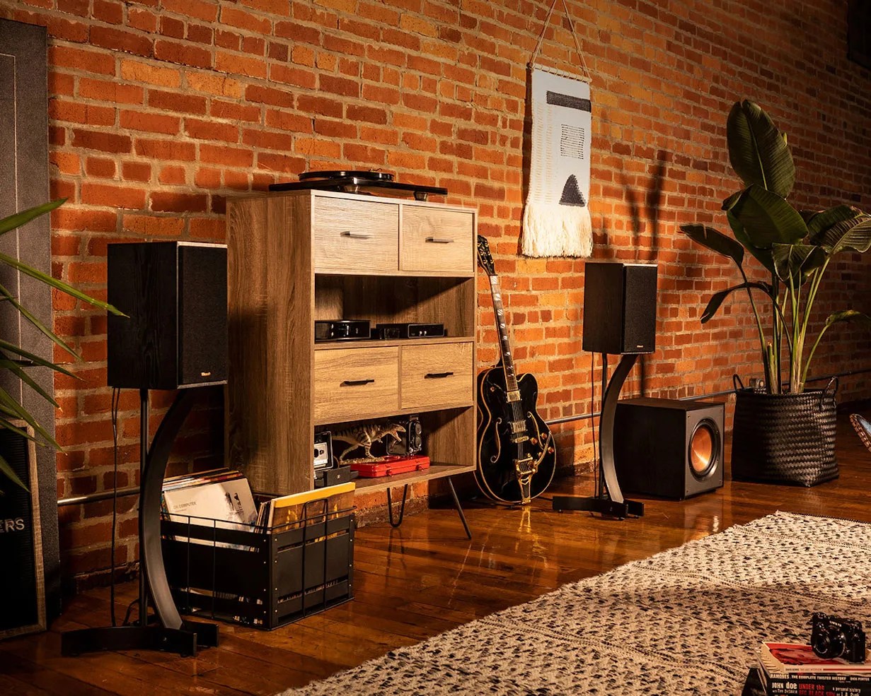 Wooden media cabinet with drawers, black guitar, black speakers on stands, and a large potted plant against a brick wall.