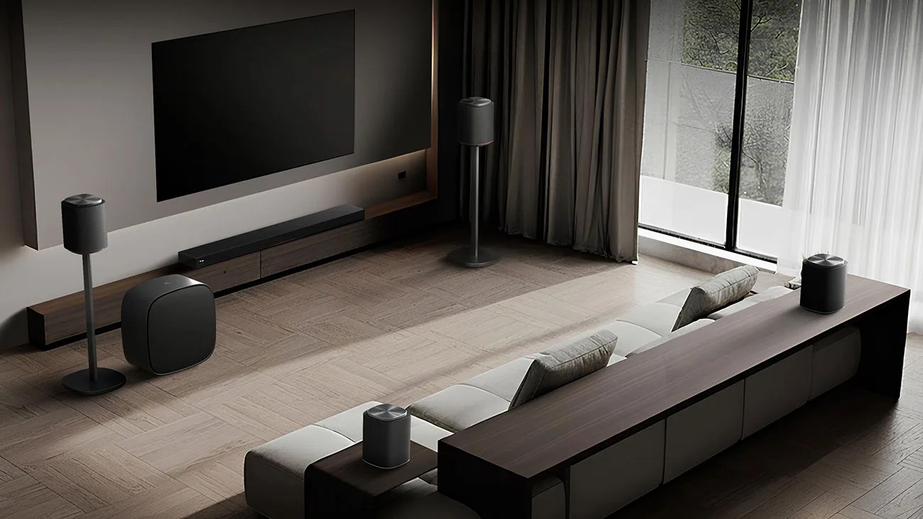 Modern living room with a large flat-screen TV, black soundbar, subwoofer, two speakers on stands, and a gray sectional sofa with a wooden console behind it.