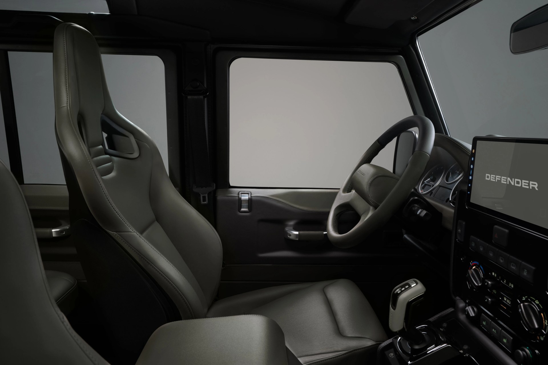 Driver-side interior of a Land Rover Defender with gray leather sport seat, steering wheel, and digital display showing "DEFENDER.