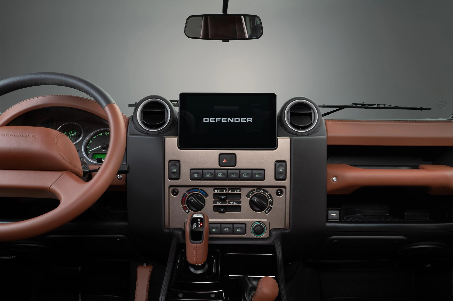 Car dashboard with tan leather steering wheel and gear shift, central touchscreen displaying "DEFENDER," and round air vents.