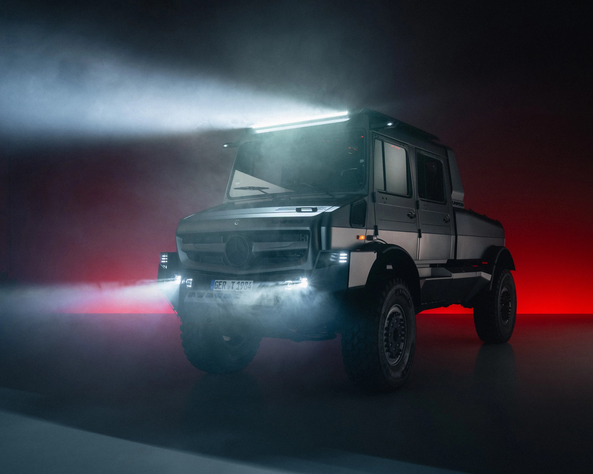 Matte gray Mercedes-Benz off-road truck with bright LED lights and fog in a dark studio with red backlighting.