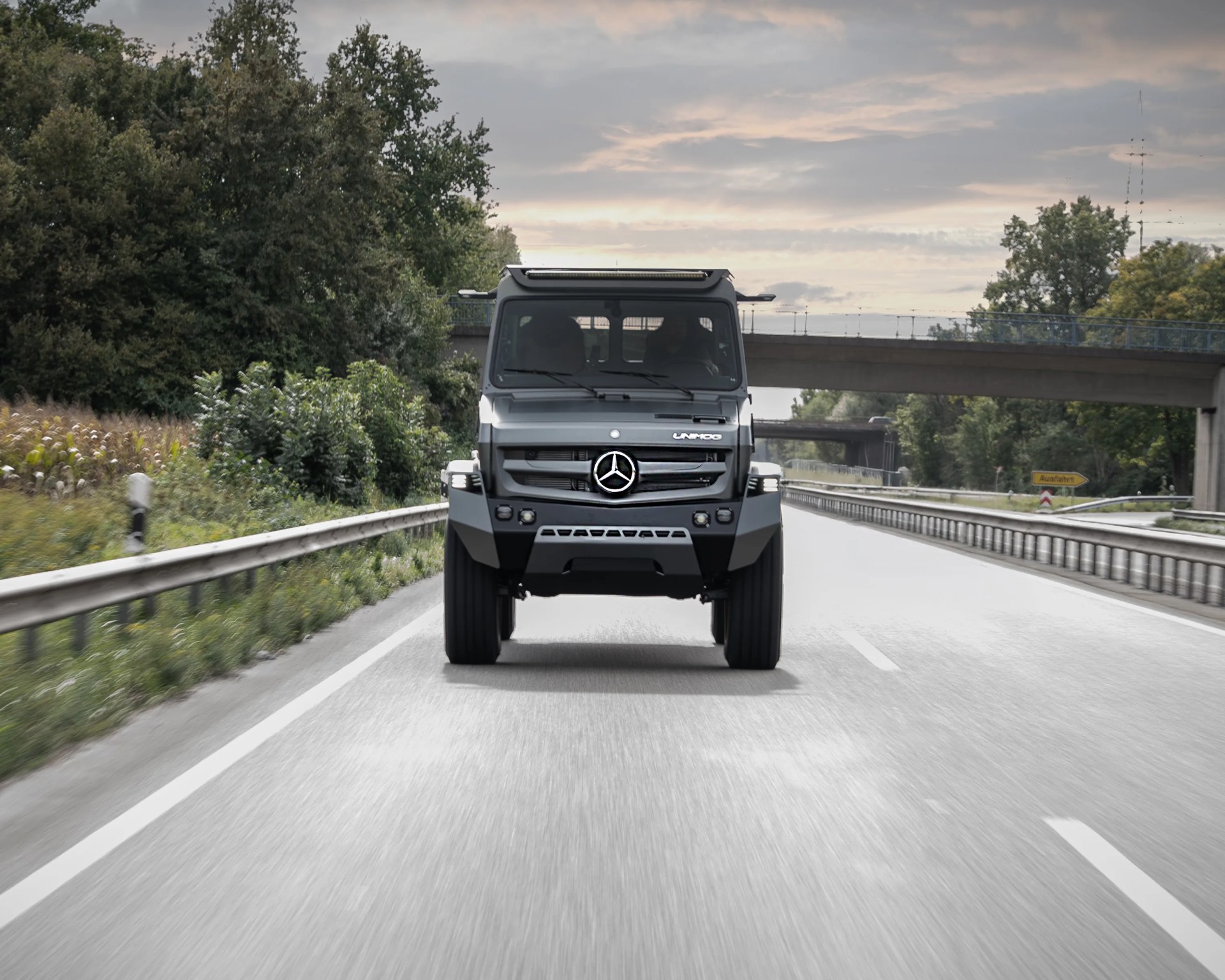 Front view of a gray Mercedes-Benz Unimog truck driving on a highway with trees and an overpass in the background.