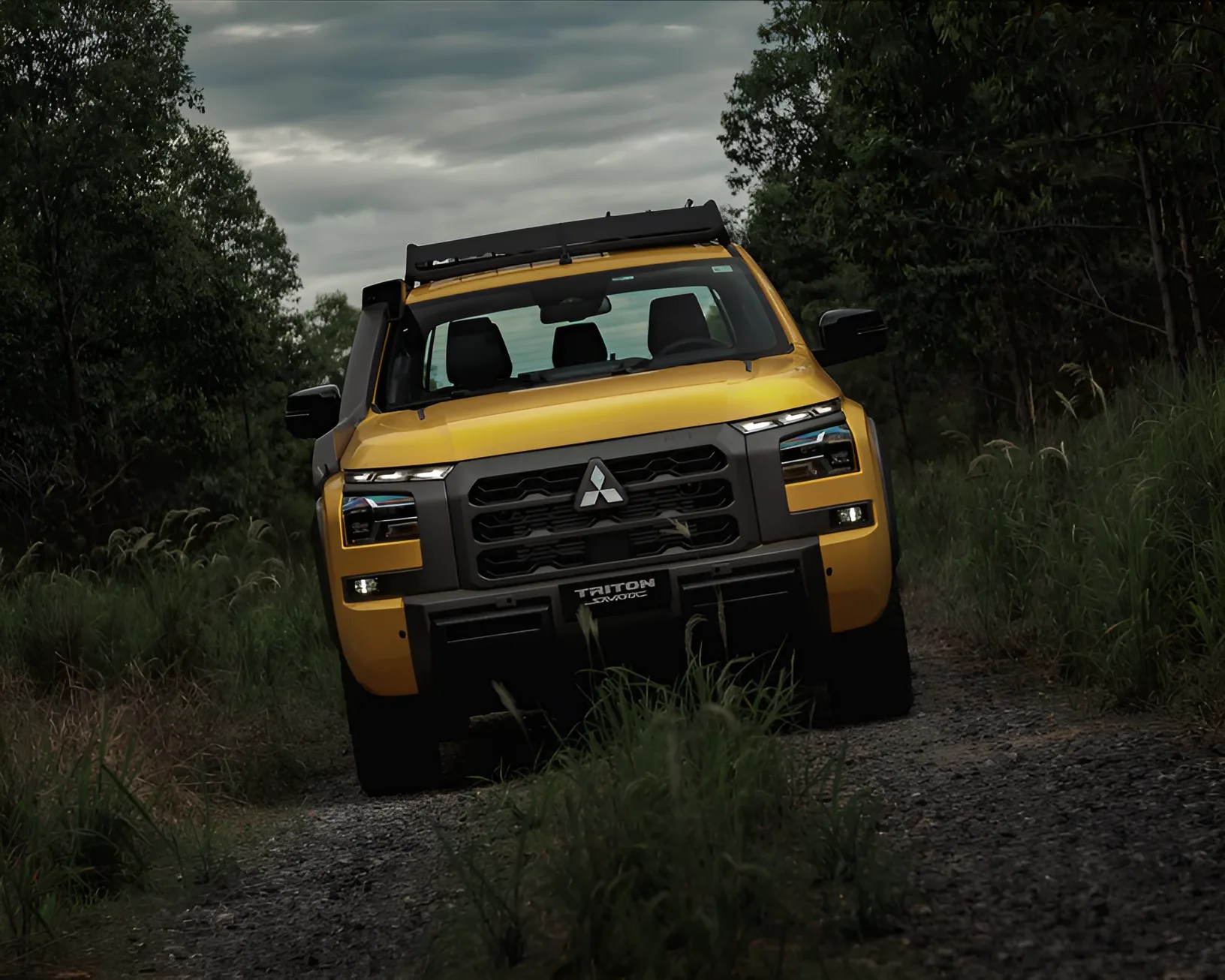 Yellow Mitsubishi Triton 4x4 pickup truck on a gravel forest path under a cloudy sky.
